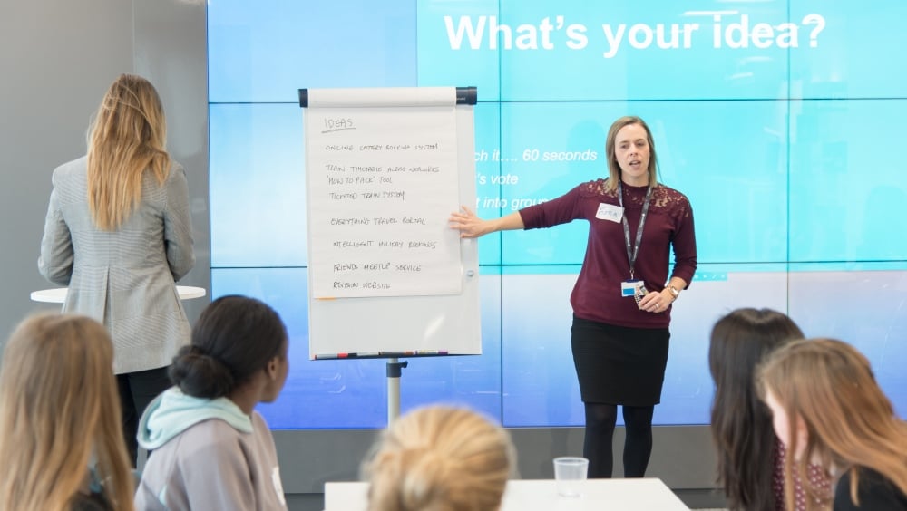 A woman speaks to a group of girls with "Ideas" written on a flipchart next to her